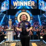 Luke Littler playing darts on stage during a professional tournament with fans watching
