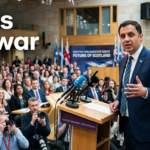Anas Sarwar speaking at a political event with a Scottish flag backdrop while addressing supporters and the media.